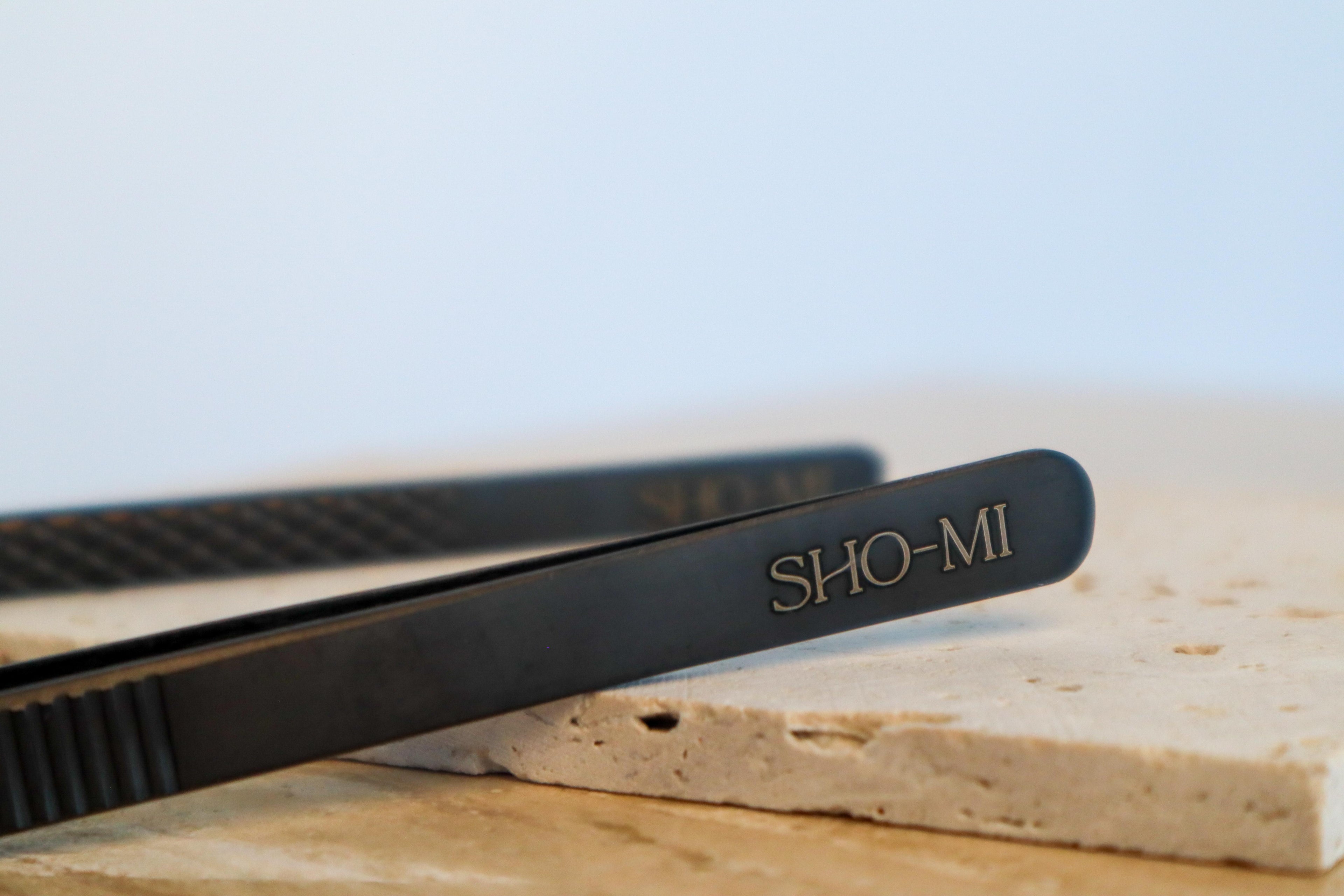 Black SHO-MI branded tweezers on a light surface with a blurred background