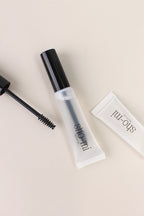 A bottle of brow gel with a black cap, alongside a brush, on a light background.